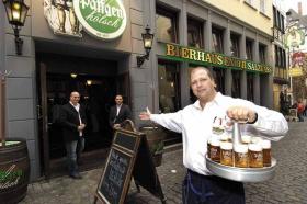 Köbes mit Kölsch-Kranz in der Hand vor dem Restaurant